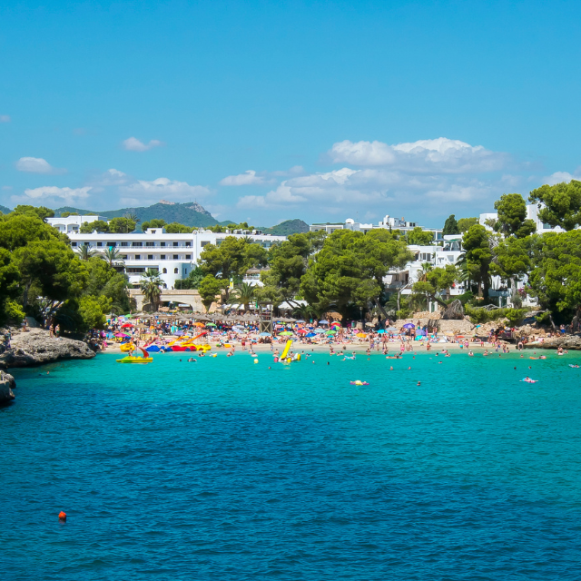 Blick vom Wasser auf den Strand voller Leute und Liegen, im Hintergrund Hotels
