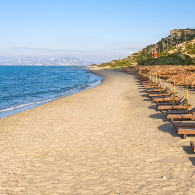 Blick entlang des hellen Sandstrands Kardamena Beach mit vielen aufgereihten Liegen und Sonnenschirmen auf der rechten Seite
