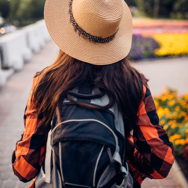 Blick von hinten auf Frau mit Rucksack und Sonnenhut, die durch Blumengarten wandert