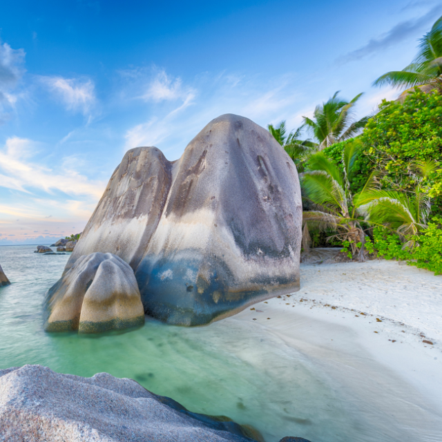 Heller Sandstrand mit Granitfelsen im Wasser und Palmen im Hintergrund