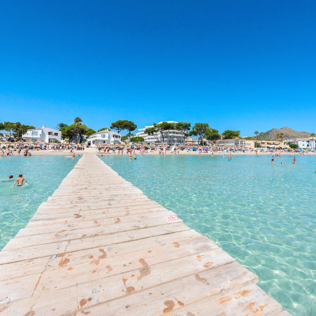 Blick vom Steg im Wasser auf den Strand voller Liegen und Schirme mit badenden Leuten