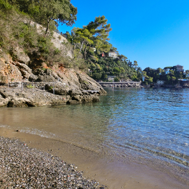 Blick vom dunklen Sand auf das blaugrüne Wasser und Holzstehe im Hintergrund an den Klippen