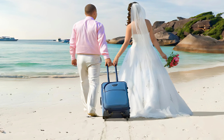 Paar in Anzug bzw. Brautkleid und kleinem Koffer laufen über Strand