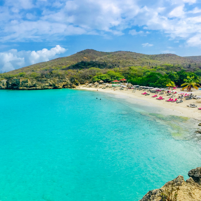 Blick von angrenzender Klippe auf die Bucht mit hellem Sandstrand, türkisem Wasser und grünen Hügeln im Hintergrund